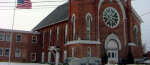 A historic red-brick church with tall arched windows and a large circular stained-glass rose window above the main entrance. A connected brick building sits to the left, and an American flag waves on a pole in front.