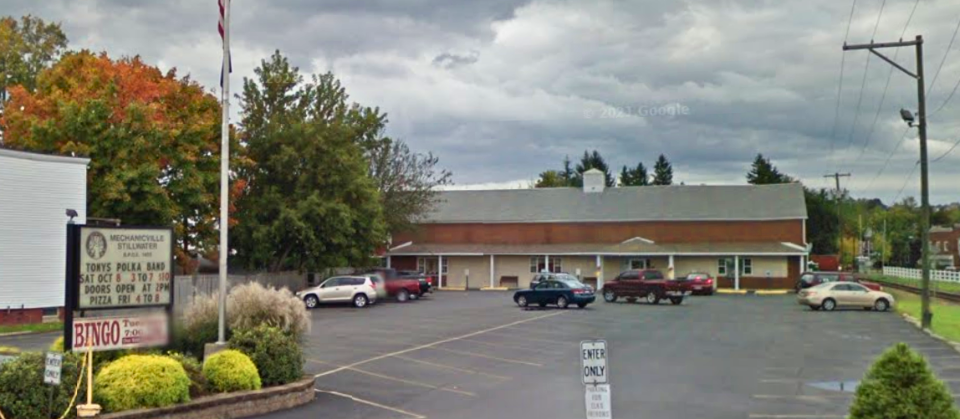 A wide view of a one-story brick and tan building with a sloped gray roof, surrounded by a large paved parking lot with several parked cars. A sign in the foreground reads “Mechanicville Stillwater Elks Lodge” with event information below it. Mature trees with early autumn foliage stand to the left, and power lines stretch across the right side under a cloudy gray sky.
