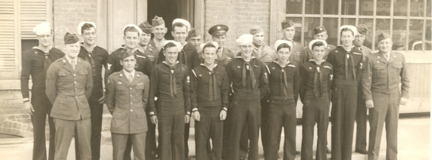 A vintage black-and-white group photograph of about twenty young service members standing in several rows outside a brick building. The group includes men in both U.S. Navy uniforms—dark jumpers with neckerchiefs and white sailor caps—and U.S. Army uniforms with garrison caps and jackets. They stand close together, smiling or maintaining neutral expressions, posed formally in front of large windows and a doorway.