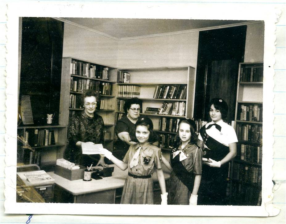 A vintage black-and-white photo of a library interior with bookshelves lining the walls. Two adult women stand behind a desk—one handing a document or paper to a young Girl Scout who stands in front of her. Three Girl Scouts, wearing uniforms with sashes and gloves, stand together in the foreground, smiling. One girl holds the paper being given to her. Library supplies, books, and a typewriter sit on the desk, and shelves of books fill the background.