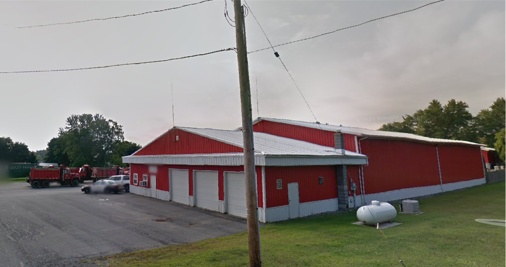 Photo of a red municipal garage building with white trim and multiple garage doors. Several trucks are parked nearby, and a utility pole stands in the foreground. The building sits beside a paved lot with grassy areas and trees in the background.