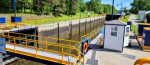A view of a canal lock system with concrete walls and bright yellow railings along blue metal walkways. A small white control building sits beside the lock, and grassy embankments and trees line the waterway.